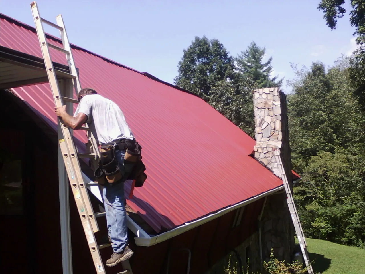 Roofer on a ladder beside a red metal roof and stone chimney — Soffit & Fascia Repair by Roofing Brothers in Mcmechen